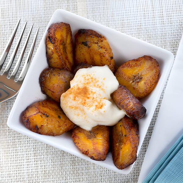 A white rectangular dish filled with golden-brown fried plantains topped with a dollop of cream and cinnamon.