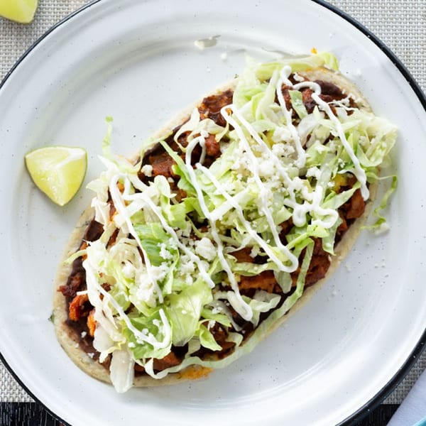 A Machete Taqueria huarache with meat, beans, lettuce, crema, and cotija cheese, with a lime wedge.