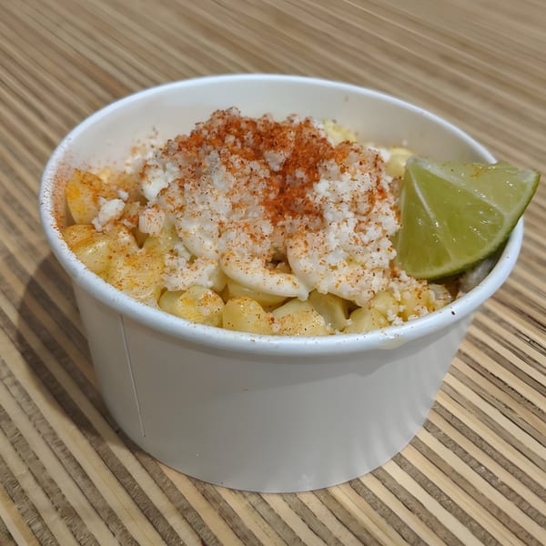 Mexican street corn (Esquites) in a white cup, topped with cheese, chili powder, and a lime wedge.