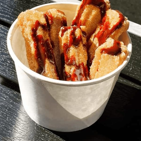 A white cup filled with churros, drizzled with a rich red sauce, on a dark textured surface.