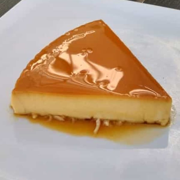 A triangular slice of flan with golden caramel sauce on a white plate.